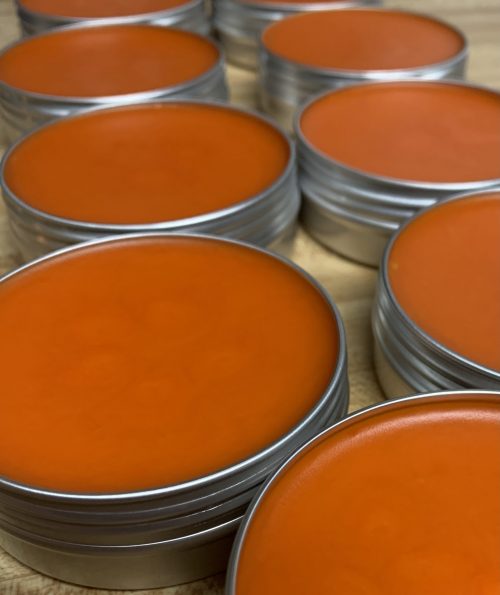 Multiple small tins containing a red-orange beeswax-based balm.