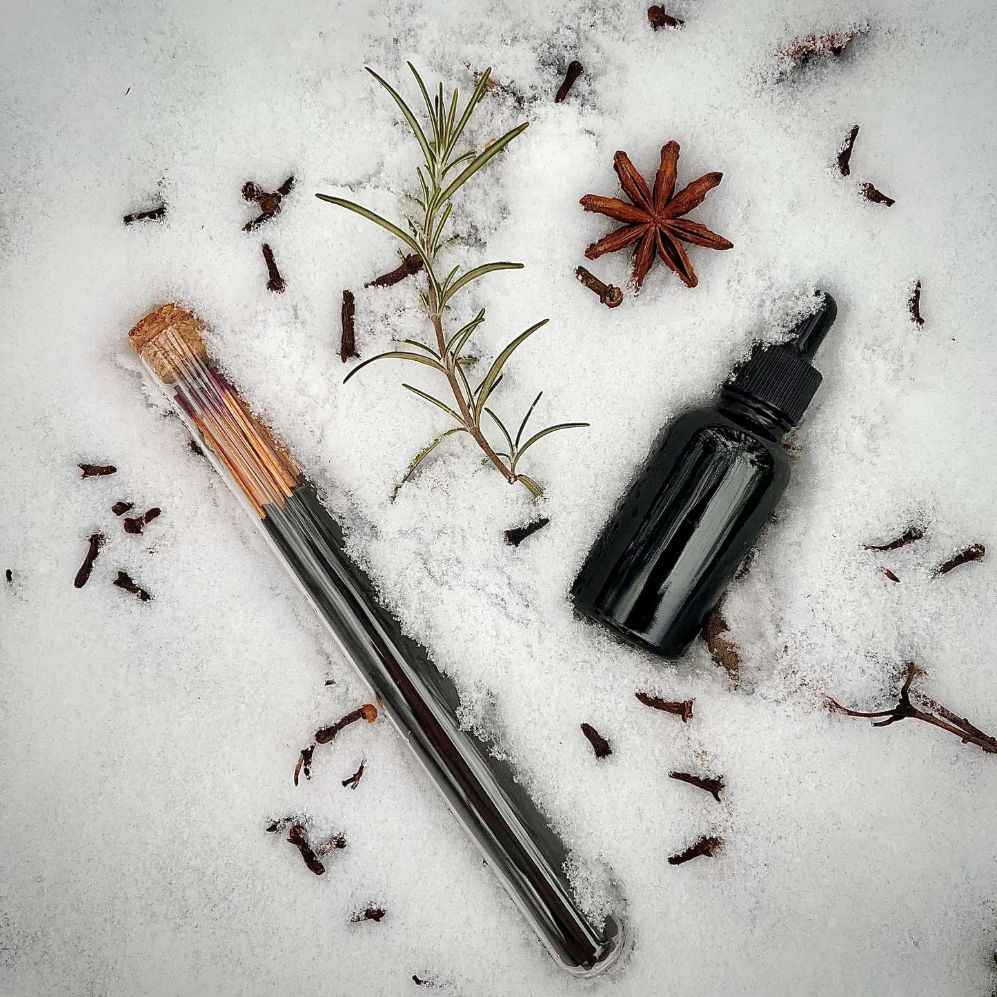 A glass tube of Sister of Mother Earth Cunning Incense rests in the snow surrounded by scattered cloves, a rosemary sprig, a star anise pod, and a dark amber brown bottle.