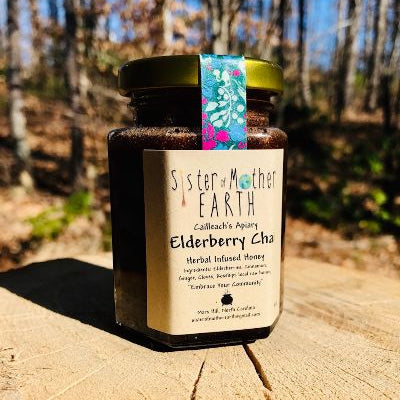 A hexagon jar of Sister of Mother Earth Elderberry Chai Infused Honey on a wooden surface with a natural background.