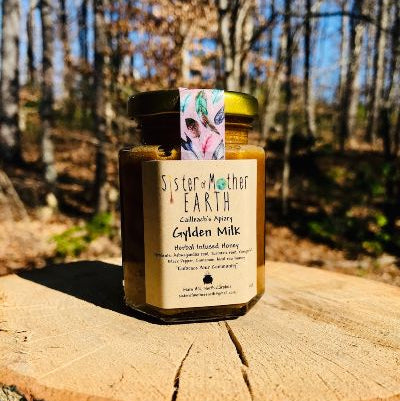 A hexagon jar of Sister of Mother Earth Gylden Milk Infused Honey resting on a wooden stump with a natural background.