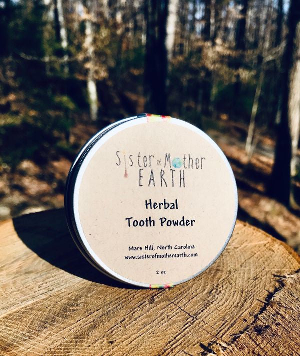 A tin of Sister of the Earth Herbal Tooth Powder placed on a wooden surface with a forest background.