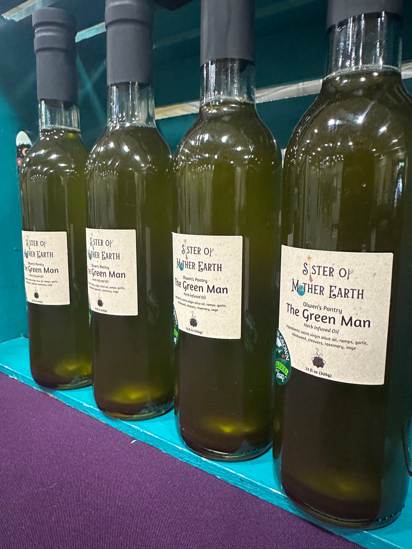 Four glass bottles of Sister of Mother Earth The Green Man Herb Infused Oil on a teal surface with a purple tablecloth. The oil is a deep olive green.