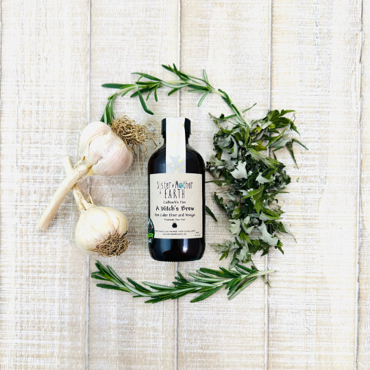 An amber glass bottle of Sister of Mother Earth A Witch's Brew Fire Cider is surrounded by garlic, mugwort, and rosemary on a light wooden background.