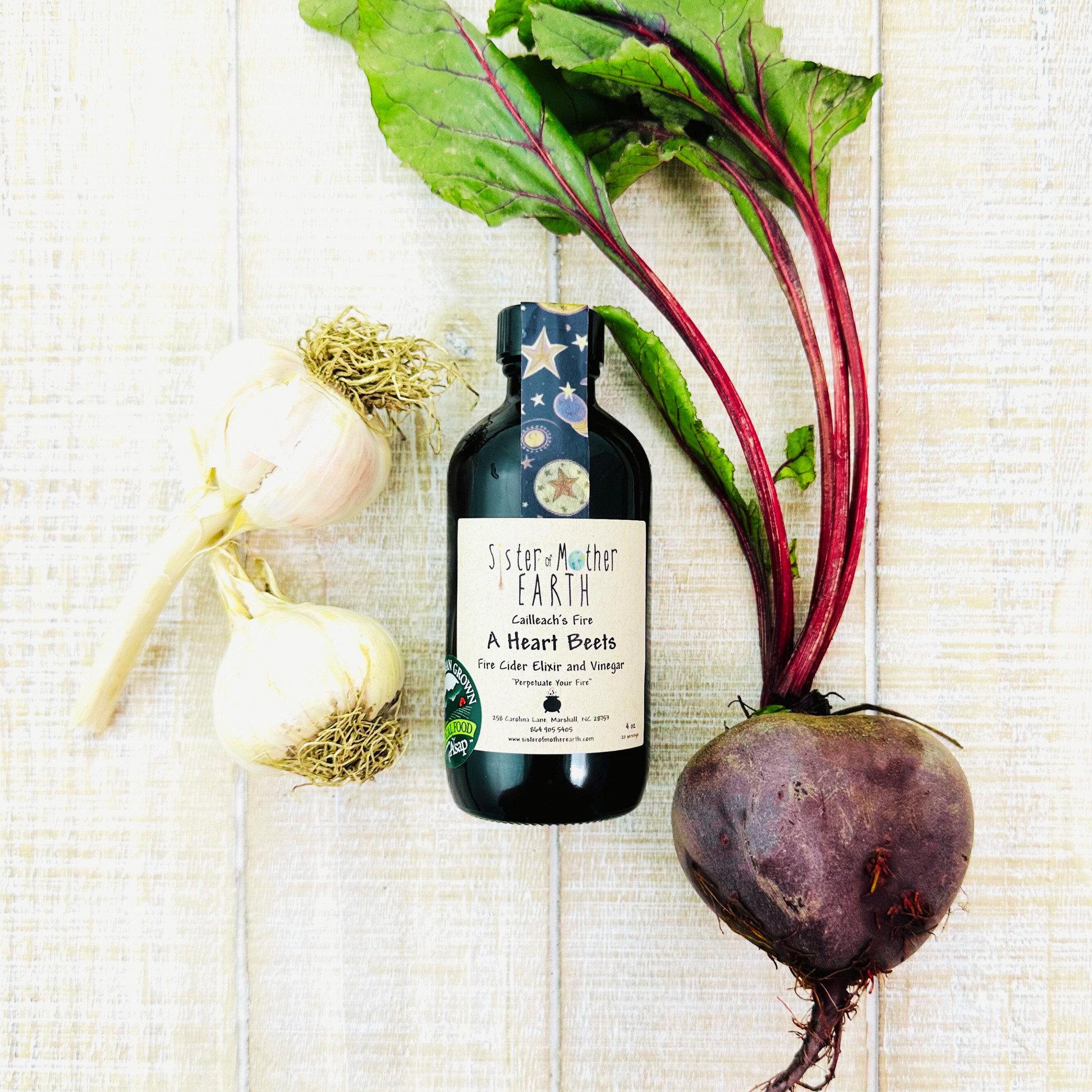 Amber glass bottle of Sister and Mother Earth A Heart Beets Fire Cider surrounded by a large beetroot and garlic on a light wooden surface.