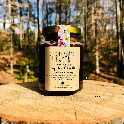 A hexagon jar of Sister of Mother Earth By Her Hearth Infused Honey placed on a wooden surface with a forest background.