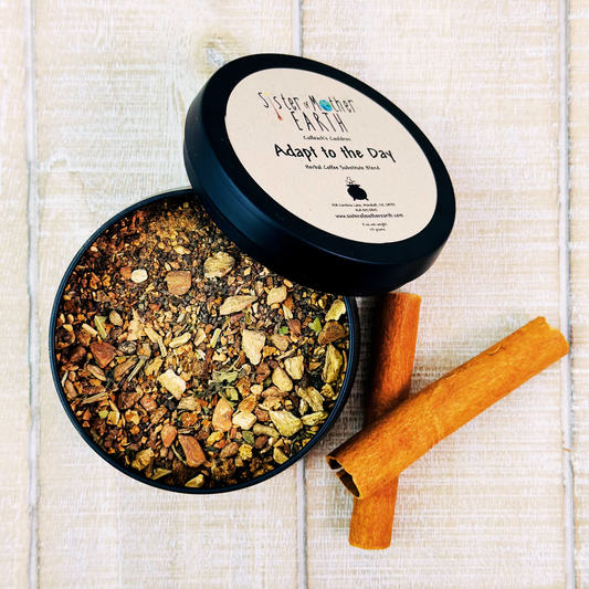 A black tin of Sister of Mother Earth Adapt to the Day Herbal Coffee substitute is open, showing brown roots and herbs, coupled with two cinnamon sticks on a light wooden surface.