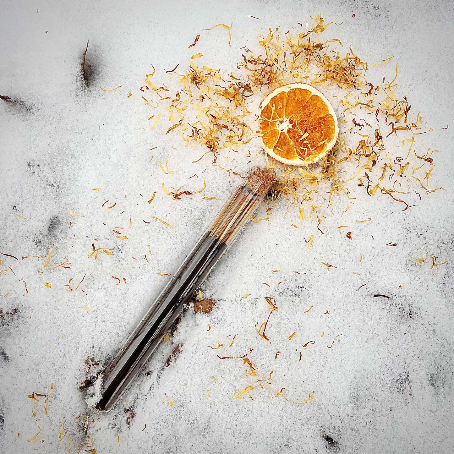 A tube of Sister of Mother Earth Sun Wakener incense lies in the snow. At the tip of the tube is a dried orange slice surrounded by golden calendula petals, giving the tube the appearance of a glowing magick wand.