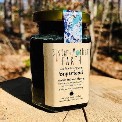 A hexagon jar of Sister of Mother Earth Superfood Infused Honey with a label that includes the product name and a brief description, placed on a wooden surface with trees in the background.