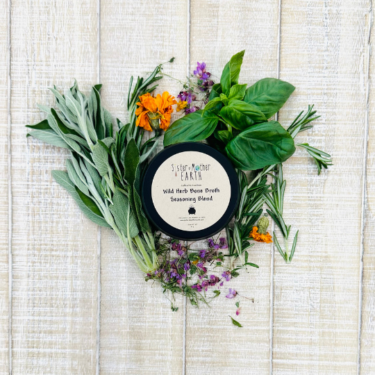 A black tin of Sister of Mother Earth Wild Herb Bone Broth Seasoning Blend surrounded by loose herbs like sage, basil, orange marigold, and rosemary on a light wooden background.